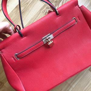 En röd handväska i (oäkta) läder med guldiga dragkedjor och smådetaljer. Axelrem medföljer. Aldrig använd. 