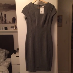  - Exklusiv, sofistikerad klänning från HM. Ganska mörk grå, med svart dragkedja som går längs hela ryggen och kan dras upp och ner hela vägen. Härligt material, man känner sig väldigt fin och proffesionell. Aldrig använd, beställd på nätet, prislappen är kvar. Köpt för 449 kr.