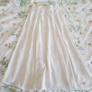 Vit långkjol med knappar - Härlig kjol med jättefint fall, 91 cm lång 😍 Köpt second hand men i jättefint skick! Saknar märkning men gissar att den passar 34-36 bra, möjligtvis en liten 38 eftersom den har resår. Möts upp i Uppsala eller Stockholm, annars står köparen för frakt 