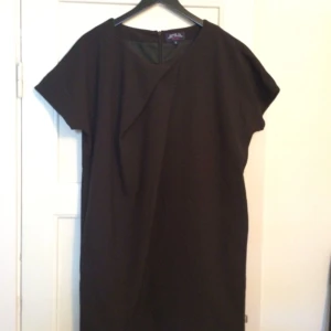  - Svart klänning i rak klassisk modell. Skönt fodrat material och fint fall, snygg draperad detalj över bröstet. Längd cirka 87 cm. Strl M. Oanvänd.  Kan mötas upp för affär eller skicka! 