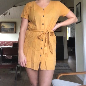 Gul klänning NAKD - Gul/senapsgul klänning från NAKD, knappar fram och band i midjan. ”Linen look buttoned dress” linneliknande material. Storlek 40.
