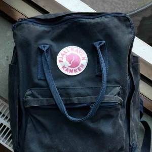  - Traditionell blå Kånken ryggsäck i ”original” storlek. Köpt för ca 5 år sedan men inte använd på flera år. I gott skick, med några slitningar undertill.   Sittunderlag ingår. Säljes pga av garderobsrensning!