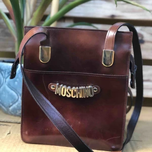  - Vintage moschino läderväska! Skulle gissa att denna är från 90/tidigt 00-tal. Relativt korta väskband vilket gör att den sitter snyggt på kroppen (på mig som är 167 går den ner till precis under midjan)
