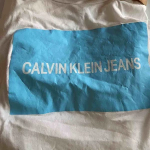  - Vit t-shirt med blått tryck från Calvin Klein! I super skick förutom lite skrynklig, vilket lätt går att fixa. Köpt för Nelly för något år sen, men knappt använd. Är storlek S men passar XS-M också beroende på önskad passform.   !! Frakt ingår ej !!