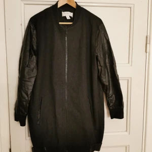 Lång Bomberjacka - Material filt och fuskläder. Spännen på vardera sida längst ner. Fickor med läderdetaljer. Text på ryggen: SILENCE. Dragkedja åt båda håll. Märke: Notion 1.3.