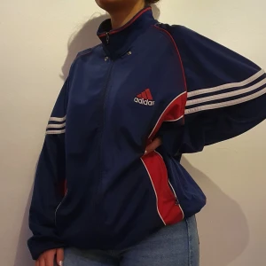 Adidas Jacka - Vintage Adidas jacka, använd väldigt sällan därför säljs jackan. Jackan är i bra skick dock kan dragkedjan vara lite seg. Stort Adidas märke på ryggen som tar upp hela ryggen. På bilderna ser ni hur jackan sitter på kille 183cm samt tjej 160cm.