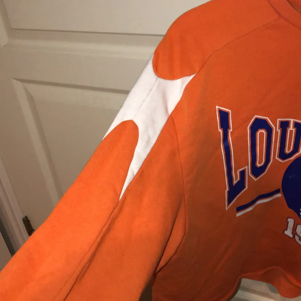  Denna tröja är orange och har vita och blå tryck samt amerikansk fotboll hjälm, denna tröja är en magtröja och har vita detaljer på armarna.. Tröjor & Koftor.