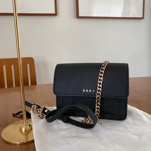 DKNY axelremsväska  - En superfin svart väska från DKNY som kan bäras på många olika sätt. Jag har använt den både över kroppen och kortat upp väskbandet och burit den över axeln. Väskan är lite sliten på kjedjsn och själva mätkrt men annars är den i fint skick. Dustbagen inåt även. Nypris ca 2500kr