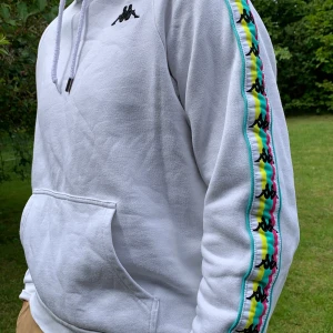 Vit kappa hoodie - Hoodie i herrstorlek S (visas på en M) med färgglada revärer på ärmarna. Använd ett fåtal gånger så är i väldigt gott skick. Dock på en av snörerna har metallgrejen ramlat av (se bild). Nypris 579kr. OBS! Buda i kommentarerna från 250kr!! Frakt tillkommer