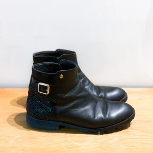 Svarta boots - Jättesköna boots i äkta läder med snyggt glansig kappa baktill. Guldiga detaljer. Dragkedja på insidan skon. Bra använt skick. Kan inte ha efter att ha opererat foten även om jag älskar dem. Stl 38.  Har Swish. Kan mötas eller skicka. Rökfritt & djurfritt hem.