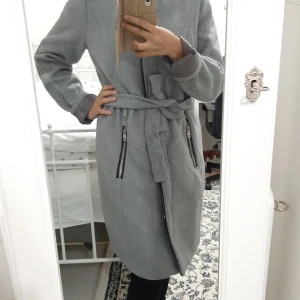Elegant & Chic Grå Höst/vinterkappa - Den här kappan är perfekt för dig som vill se elegant ut nu i höst! Fint silver dragkedjor på sidorna och snyggt skärpe till. Den passar dig som gillar vintage och chic stil.  Är i nyskick och har används enstaka gånger. Kan hämtas i Stockholms området.