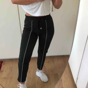  - Svarta byxor med vit ”linje” på framsidan, vita linjen är en tygbit som är påsydd, sticker ut från byxan. Mjukt material, med snören i midjan, inga fickor, passar också till en storlek S