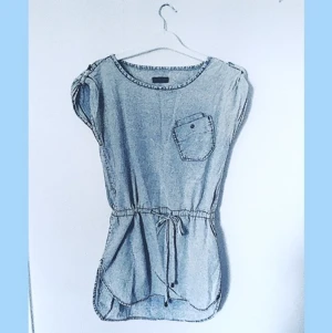 Ljusblå Klänning från ONLY - Strl 34 - Ljusblå denim(artad) klänning från ONLY i storlek 34. Har en snörning runt midjan att knyta enligt önskad passform, samt en fick på vänster bröst. Ett cirka 20cm långt snitt längst ner på vardera yttre lårben.