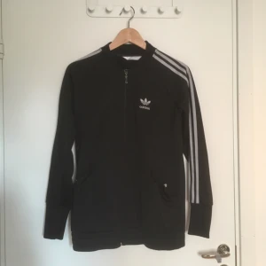  - Svart track jacket från adidas med silvriga ränder och logga. Den har mest hängt i min garderob de senaste åren och bild 3 visar det lilla slitage som finns. Annars är den i fint skick!