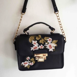 Väska - Svart handväska i måtten 19x14x6 cm. Väskan har ett litet innerfack med en dragkedja. Framtill är det broderade blommor och ett guldspänne (som är pyttelite slitet) Skriv till mig vid intresse eller om du har några frågor😄 Fraktkostnad tillkommer!  
