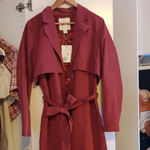 Kappa från Monki - Kappa från Monki, storlek M. Fin mörkare lilaröd färg. Tyget är matt utanpå och glansigt inut, det finns ett bälte att knyta runt midjan eller baktill. Helt oanvänd. Köpt för 600kr men säljer för 150. Perfekt till våren! Frakt tillkommer