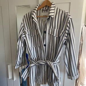 Monki randig utility jacka - Randig jacka med knappar från Monki, perfekt för tidig sommar. Jackan har fickor och ett tjockt band runt midjan. Den är i storlek XXS men passar större storlekar såsom XS och S. Den är använd ett fåtal gånger. Köparen står för frakten. 