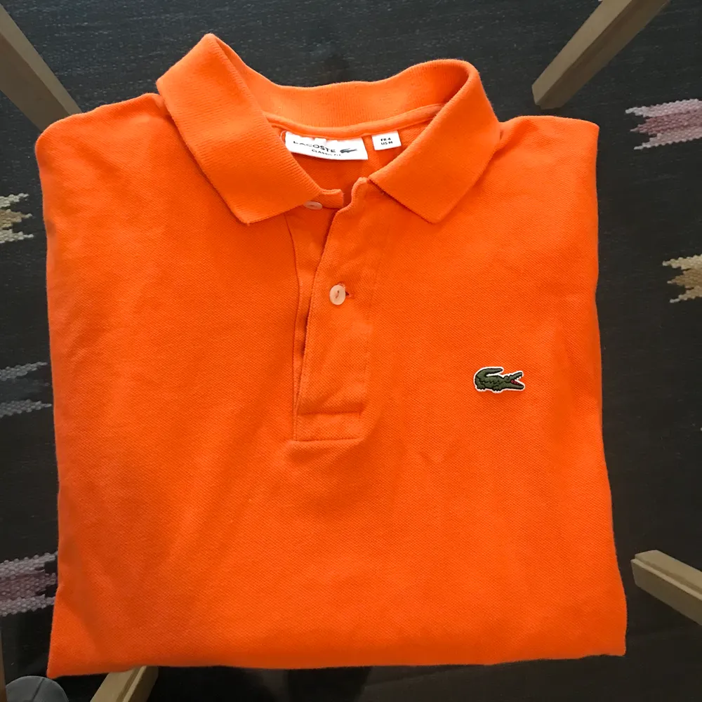 Det här är en piké från Lacoste som har en broderad logotyp på bröstet i form av en krokodil. Ärmsluten har ribbade muddar runtom. Passformen är classic fit Pikén har en knappslå med två knappar. Broderad logotyp - Knappslå med två knappar - Mjukt pikétyg - 100% Bomull - 40° Maskintvätt. T-shirts.