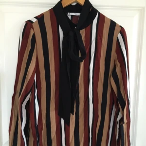  - Snygg randig knytblus i svart, brunt, vitt och beige. Skön kvalitet, använd en gång. Dold knäppning fram, små diskreta guldknappar på manschetter och överst vid halsen. 