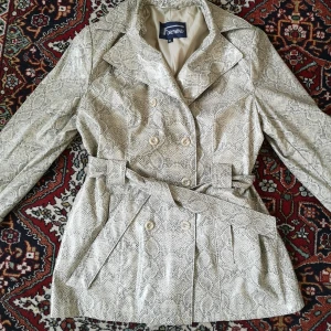 Fake läder jacka/kappa i ormskinn - Jacka/kappa i fake läder/lack . Dubbelknäppt med skärp i midjan. Står stl L i den, men sitter snyggt oversize på mig som är en S. 