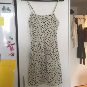 Rosettklänning stl 38 - Vit klänning med svarta rosetter på. Två fickor i kjolen, knappar fram och spagettistraps som går att göra minsta och större 