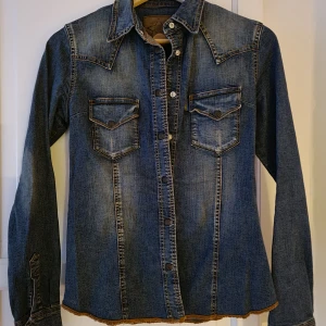 Gul&blå jeansskjorta - En supersnygg jeansskjorta av kända märket Gul&Blå i den perfekta färgen denimblå. Lite stretchig I materialet och har markerad midja. Kända detaljer på så som sömmen och märket på fickan finns tydliga samt tryckknappar. Strl S. Helt ny. Oanvänd. Inga defekter. Köpt för 799,-  säljer för 399,- Hämtas upp el så står köparen för frakt. 