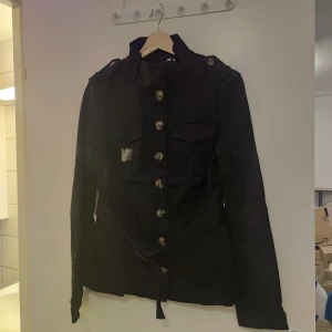 Helt ny denimjacka med bälte och knappar - Helt ny svart denimjacka, inte ens provad. Bälte med snyggt spänne, stora knappar med en "gömd" rivet-knapp högst upp. Bröstfickor. Storlek 12 eller 40 beroende på system. Ca 38-40 cm bred i midjan (när det ligger platt), 67 cm hög(platt), 50 cm ärmar. 