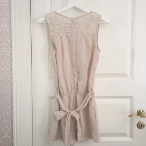  - Beige shortsdress med band, stängs med knapar, spets upptill och fickor på sidorna. 