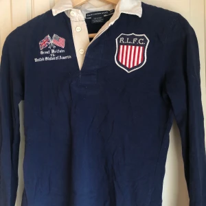 Tröja Ralph Lauren - Marinblå tröja med vit krage och broderade märken på bröstet. Från Ralph Lauren. I nyskick, aldrig använd.