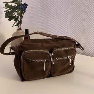 Noella väska  - Väska från Noella i mörkbrunt med silverdetaljer. Sparsamt använd. Inköpt för 699kr. Saknar det långa bandet..