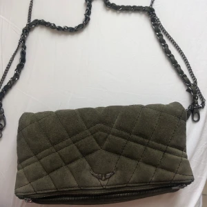 Zadig & Voltaire Clutch - Snygg väska från Zadig & Voltaire i mörkgrön mocka. Begagnat skick, med en av fästena för kedjan dessvärre trasigt. Därav priset! Har använt den som Clutch men går enkelt att laga. Kedjan kommer med. Nypris ca 4000 kr 