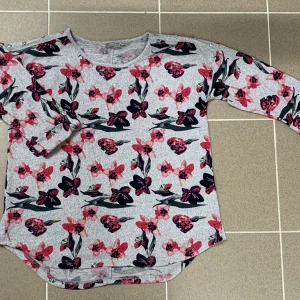 Jätte söt grå tröja med rosa blommor på! - Jätte söt grå tröja med rosa blommor på! Med knappar längst axeln, och uppvikt med knapp i slutet av ärmen. Aldrig använd endast testad! 