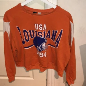 En orange magtröja med tryck -  Denna tröja är orange och har vita och blå tryck samt amerikansk fotboll hjälm, denna tröja är en magtröja och har vita detaljer på armarna.