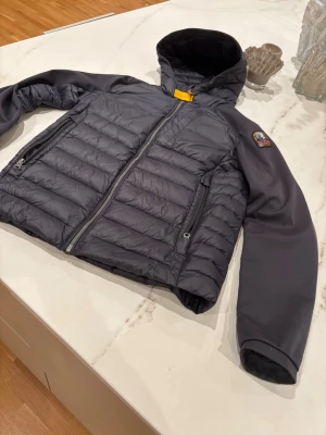 Parajumpers jacka - Snygg grå dunjacka från Parajumpers med huva och dragkedja framtill. Jackan har quiltat mönster, två sidofickor med dragkedja och en gul detalj vid halsen. Perfekt för kalla vinterdagar. Fint skick utom defekten som syns på sista bilden.