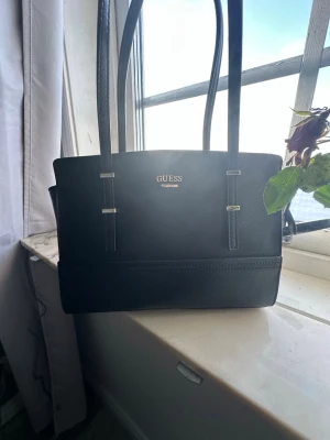 Svart handväska från Guess - Stilren svart handväska från Guess med två långa axelband och guldfärgade detaljer. Väskan har en rektangulär form och är tillverkad i ett strukturerat konstläder. Guess-loggan syns framtill i guld. Perfekt för dig som gillar klassisk och enkel design.