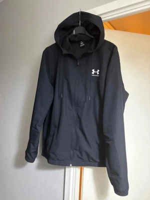 Svart hoodie från Under Armour - Säljer en svart hoodie från Under Armour med dragkedja framtill och justerbar huva med snören. Hoodien har logga i vitt på bröstet och är tillverkad i ett mjukt material som passar perfekt till chill eller träning. Klassisk och stilren design med långa ärmar.