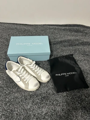 Vita Philippe Model sneakers med grå detaljer - Snygga vita sneakers från Philippe Model med grå mockadetaljer och blå häl. Klassisk låg modell med vita skosnören och diskret logga på sidan. Skorna är i läder och mocka, perfekta för en stilren och trendig look.