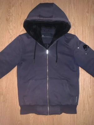 Mörkblå hoodiejacka Moose Knuckles Classic Bunny - Snygg mörkblå hoodiejacka från Moose Knuckles med dragkedja framtill och stor huva. Jackan har mjukt foder i pälsimitation och quiltad insida. Praktiska fickor framtill och logga på ärmen. Perfekt för kyliga dagar. I bra skick men finns tecken på användning, lite solblekt i färgen samt små minimala slitningar vid fickorna. Storlek XS men passar en S bättre 