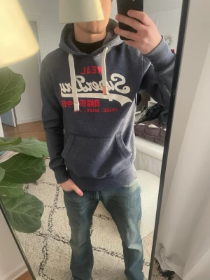 Mörkblå Superdry hoodie med tryck - Snygg mörkblå hoodie från Superdry med vit och röd logga på bröstet och vita snören i huvan. Stor ficka framtill och trycket '34' på ena ärmen. Mjuk bomullsblandning och klassisk passform, perfekt för en avslappnad streetwear-look.