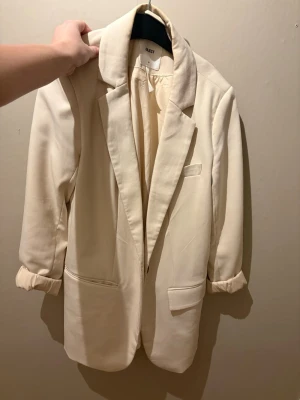 Krämvit kavaj från Object - Stilren krämvit kavaj från Object med klassisk passform, slag och två framfickor. Ärmarna är uppvikta för en avslappnad look och materialet känns mjukt och följsamt. Perfekt för dig som vill ha en enkel men snygg kavaj till garderoben.