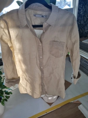 Beige linneskjorta med ficka - Snygg beige skjorta i linne med klassisk krage och bröstficka. Skjortan har långa ärmar som kan vikas upp och knäppning framtill med vita knappar. Perfekt för en avslappnad men stilren look.
