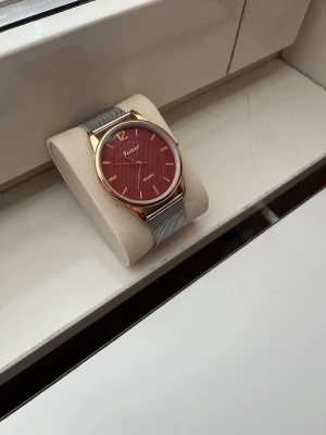 Röd klocka med meshband från Pascalio - Stilren klocka från Pascalio som har används en gång. Denna modell har en vinröd urtavla och guldfärgat boett. Klockan har ett silverfärgat armband som ger en modern look. Urtavlan har minimalistiska guldmarkeringar och ett diskret mönster i mitten. Perfekt accessoar för att lyfta din outfit. Nypris cirka 1200kr💸. Kom gärna med bud 🤝