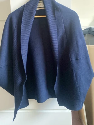 Mörkblå öppen kofta från Soft Goat - Mysig mörkblå öppen kofta från Soft Goat i ett mjukt stickat material. Koftan har en avslappnad passform med breda slag och faller snyggt över axlarna. Perfekt att slänga över en t-shirt eller topp för en chill look.