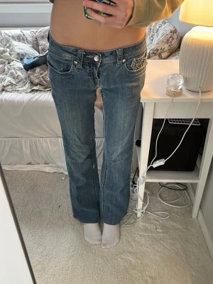 Blå bootcut jeans med pärldetaljer - Säljer ett par blå bootcut jeans med låg midja och snygga pärldetaljer samt broderier på bakfickorna. 37cm raktöver midjan. Längden passar mig som är 158.