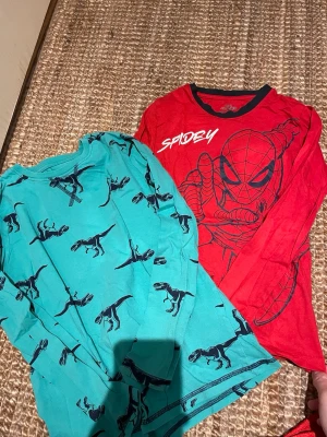 Marvel Spiderman & Dino långärmade t-shirts - Två långärmade t-shirts, en röd Marvel Spiderman med svart tryck och kontrastkrage, samt en turkos med svarta dinosaurier. Perfekta för dig som gillar superhjältar och coola prints. Båda i mjuk bomull och passar barn 6-8 år.
