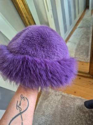 Lila fluffig pälsmössa - Säljer en supermjuk och fluffig lila pälsmössa med bred pälskant runt om. Mössan har en rund form och är perfekt för dig som vill sticka ut med en färgstark accessoar. Materialet känns lyxigt och ger en cool vintagekänsla.