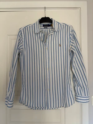Randig skjorta från Polo Ralph Lauren - Klassisk vit skjorta med blå ränder från Polo Ralph Lauren. Skjortan har lång ärm, knappar framtill och en broderad logga på bröstet. En riktigt snygg skjorta från Ralph Lauren som tyvärr blivit för liten. Väldigt fin på sommaren och passar perfekt till allt!! Nypris ca 1700kr