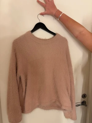 Rosa fluffig stickad tröja - Mysig rosa stickad tröja med fluffig yta och rund halsringning. Tröjan har långa ärmar och en avslappnad passform, perfekt för kalla dagar. Materialet känns mjukt och skönt mot huden. Enkel och stilren design som är lätt att matcha.