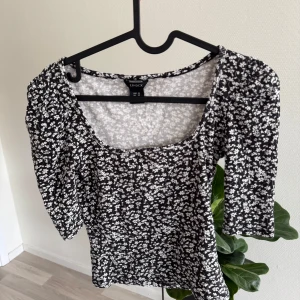 Svart blommig topp från Lindex XS - Supersöt svart topp från Lindex i storlek XS med vita småblommor över hela. Toppen har fyrkantig ringning, puffiga axlar och trekvartsärm. Den är figurnära och ribbad i materialet, vilket gör den både bekväm och snygg.