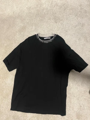 Svart t-shirt från Acne Studios - Svart t-shirt från Acne Studios med kontrasterande grå ribbad krage där varumärkets namn är invävt runt hela halsen. Klassisk passform och korta ärmar, perfekt till jeans eller shorts.